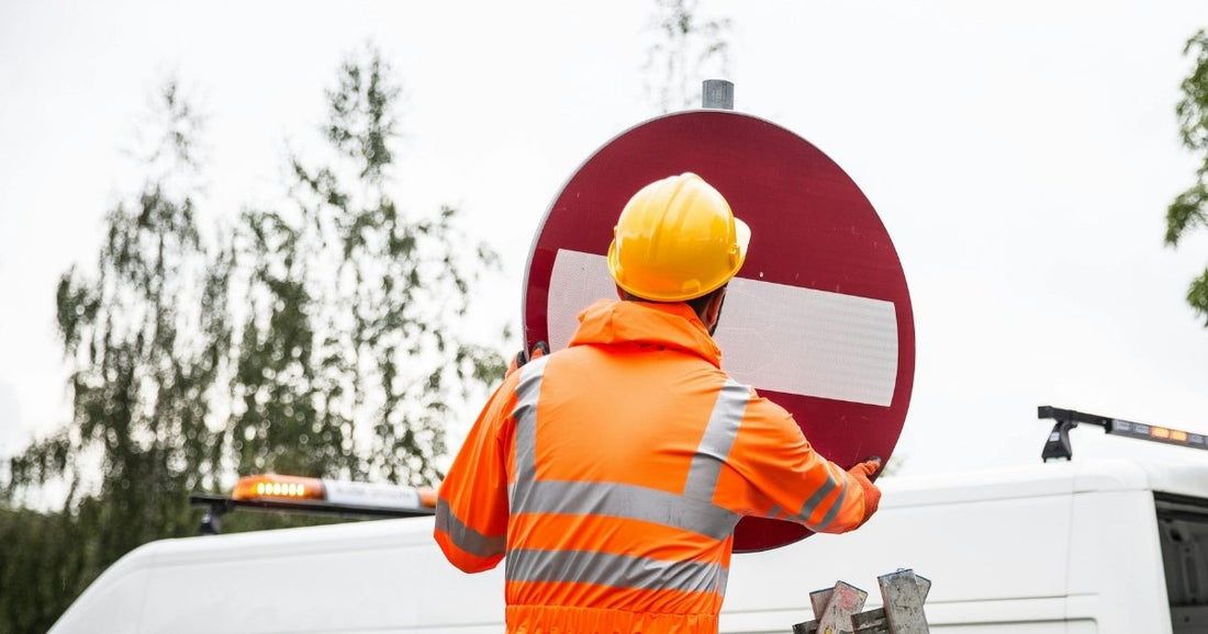 Worker in a high-visibility orange jacket and yellow hard hat installing a circular red and white road sign.